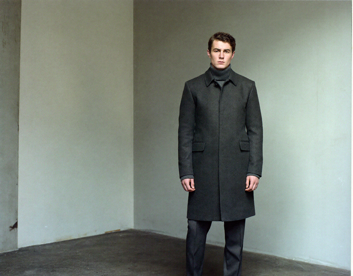Man in a charcoal wool overcoat and roll-neck in a bare concrete interior