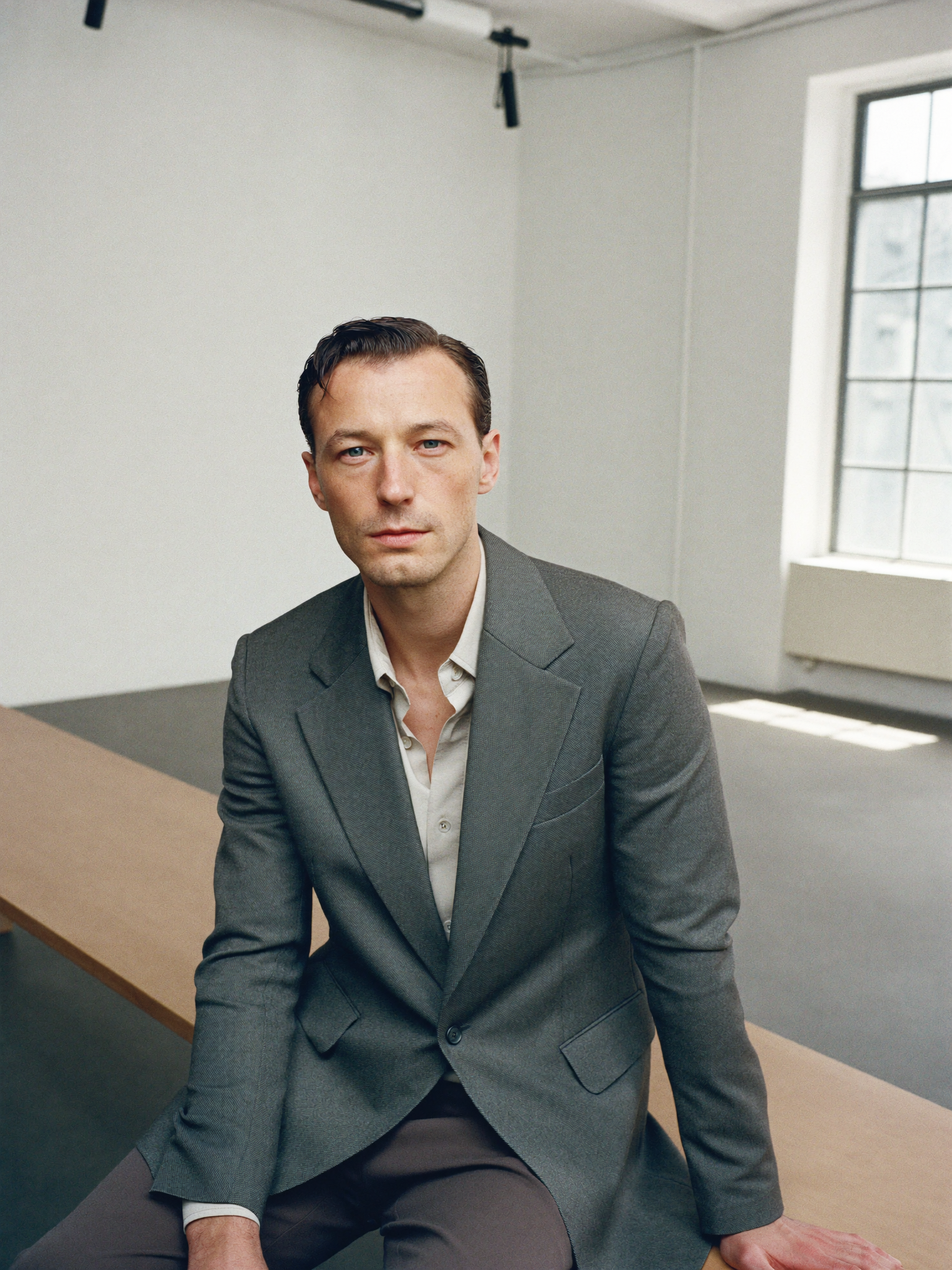 Man in a stone-grey blazer and shirt seated in a minimal studio